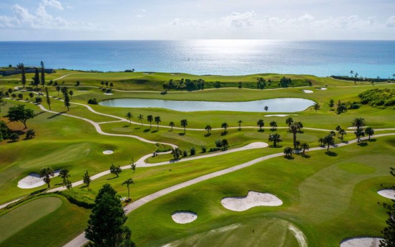 A scenic view of the Port Royal Golf Course in Bermuda, showcasing lush green fairways, sand bunkers, and the ocean in the background under a partly cloudy sky.