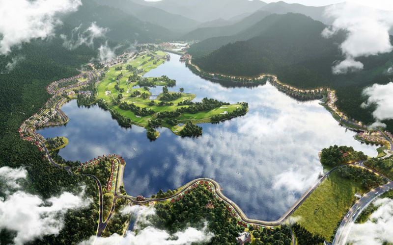 An aerial view of Thanh Lanh Valley Golf Resort featuring a lush green golf course surrounded by a large reflective lake, rolling forested hills and scattered clouds.